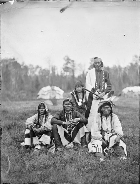 Group of five Native Americans with three men sitting, and one man and child standing. The standing man is holding what may be a pipe.