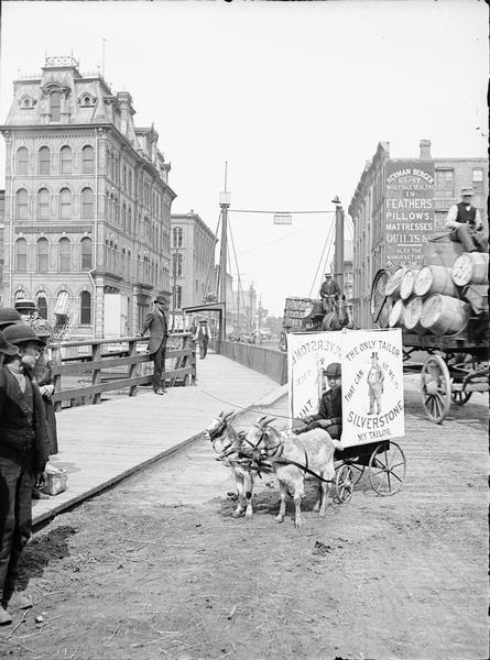 A boy on a Milwaukee street is driving a goat cart with an advertisement for Silverstone the tailor. Several on-lookers are watching from the left hand side of the street. Behind the boy in the goat cart are two loaded wagons on a bridge.