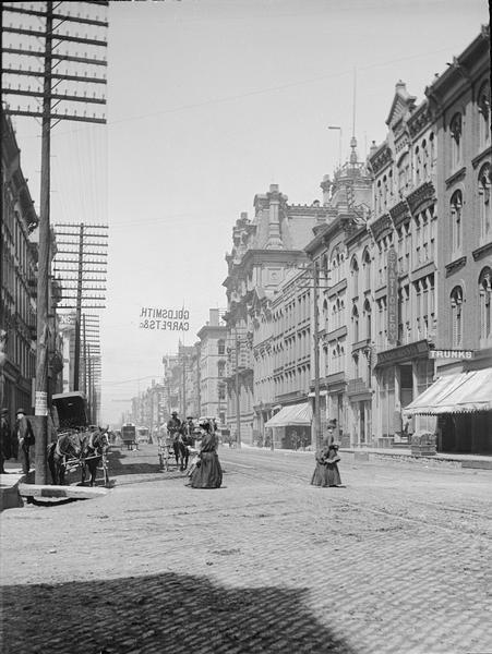 East Water Street looking North from Huron Street. There is an advertisement over street for Goldsmith Carpets. Women are crossing the street.