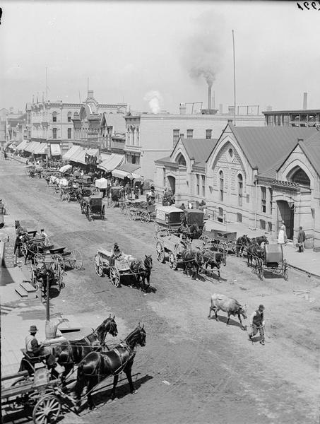 Elevated view of a farmer's market with horse-drawn vehicles. One man is leading a bull down the street.