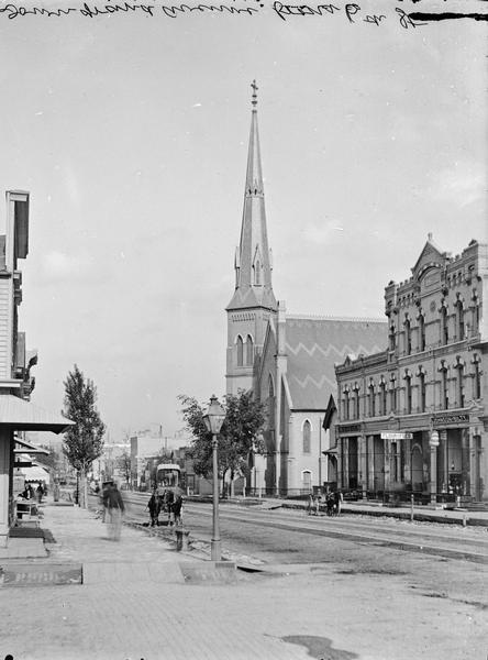 View down Grand Avenue from 6th Street. On the street are several horse-drawn vehicles. There is a church in the background.