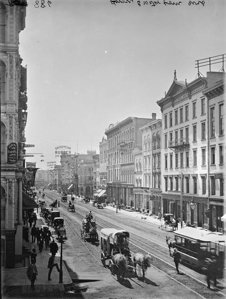 Elevated view down East Water Street from Wisconsin Avenue, with buildings, people, and street traffic including horse-drawn vehicles and a horse-drawn streetcar.
