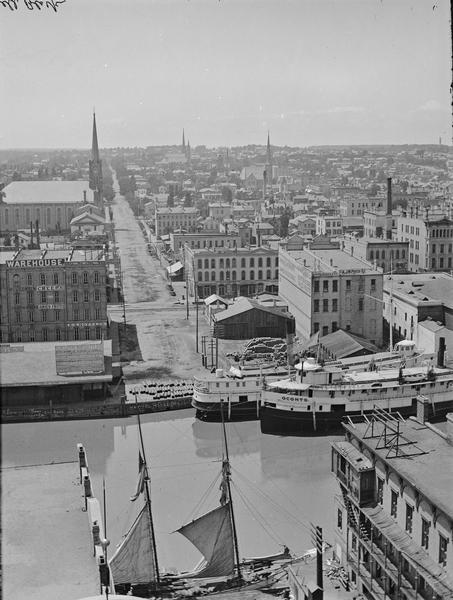 Elevated view west from Mitchell Block of warehouses, industrial buildings and a church in the background. In the foreground is a waterway with a schooner and two ships, one of which is the Oconto.