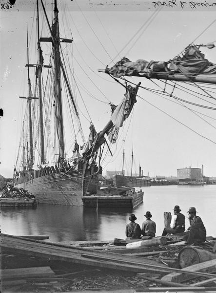 Four men are sitting on the shore and looking out at a ship on the water at the Wolf and Davidson ship yard.