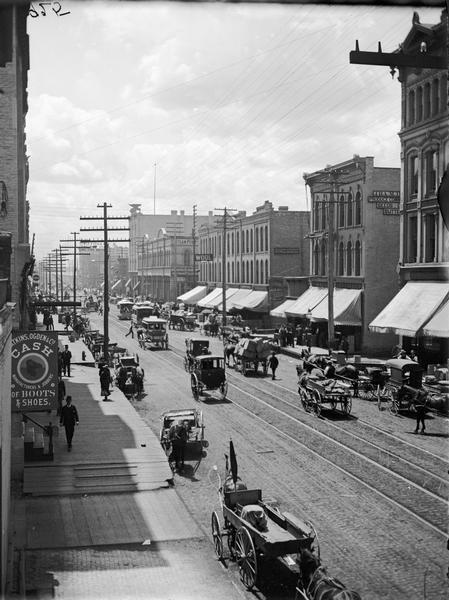 Elevated view of West Water Street from Grand Avenue of horse-drawn vehicles, pedestrians, buildings, storefronts and power lines.