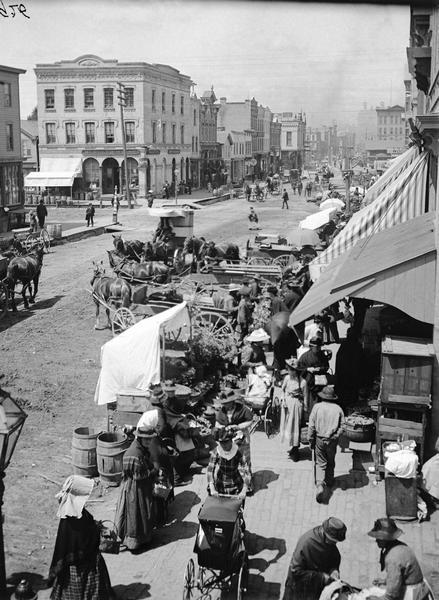 Elevated view of the Division Street Market. Men and women with baskets and baby carriages are walking along the sidewalk. Horse-drawn vehicles are parked at the edge of the street.