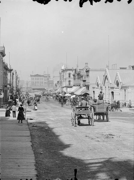 German Market on the right side of the street in the distance. Two horse-drawn vehicles in the foreground are heading toward the market. Pedestrians are walking on the wooden sidewalk on the left side of the street.