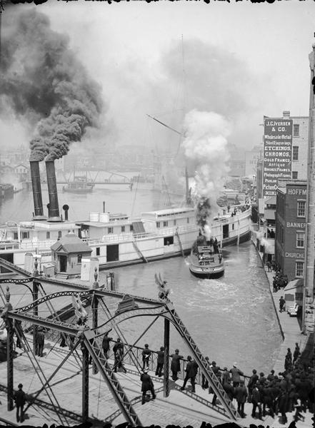 Elevated view of a Milwaukee River scene showing people watching several steamboats from a bridge.