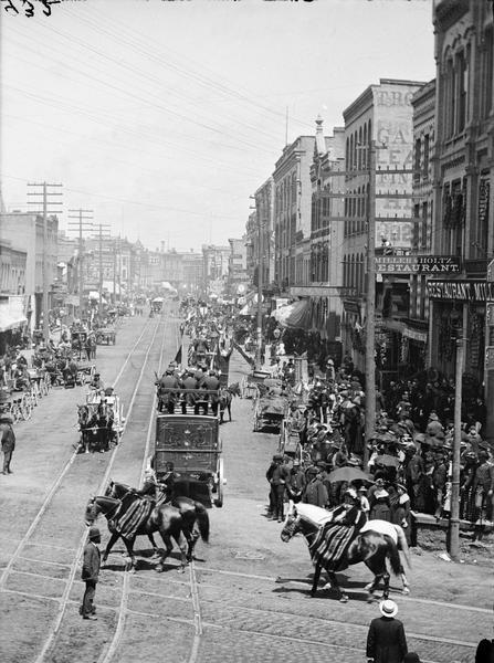 Elevated view of crowds watching a circus parade at the corner of West Water and Grand Avenue. In the foreground are four people on horseback. Beyond them is a circus wagon bearing musicians that is moving away from the camera.