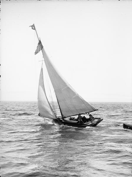 Several people are standing aboard a sailboat sailing in the harbor. A small boat is tied to the back of the sailboat.