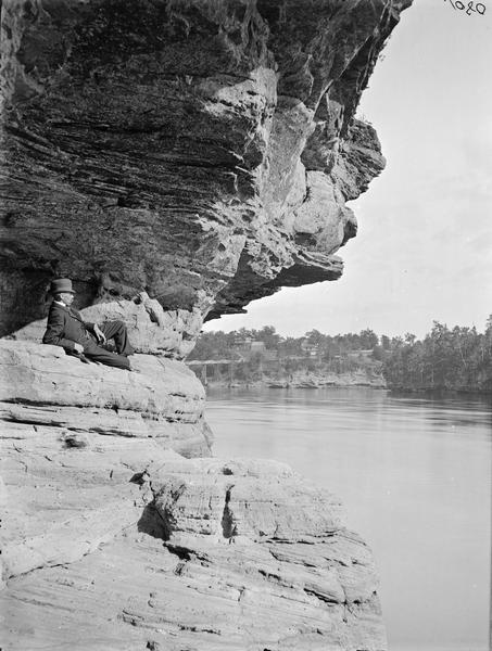 Man in dark suit and hat reclining on Angel Rock and looking down the river. There is a bridge in the distance.