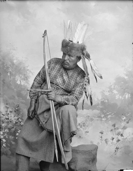 Studio portrait in front of a painted backdrop of Ho-Chunk Big Bear, holding a bow and arrow and wearing a fur hat with feathers.