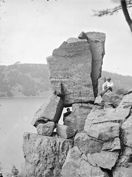 Devil's Lake Vicinity; Cleft Rock on East Bluff. There is a man sitting on the rocks on the right. There is a second man peering through a rock gap at the left.