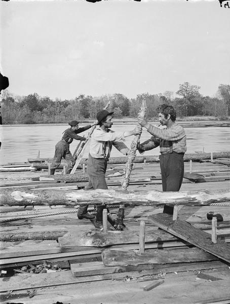 Stereograph of three men aboard a logging raft using log poles to "put down the grouzers."