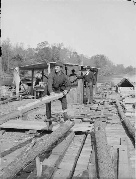 Stereograph of a  man on a raft holding the end of a hand hewn log, probably an oar, while three men watch in the background.  Two of the men are standing under a roofed shelter.