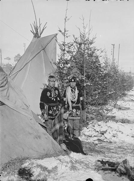 A Sioux woman is sitting on the ground, and two Sioux men are standing in front of tepees.