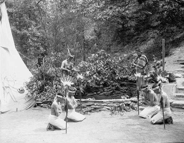Ho-Chunk flute ceremony or prayer for rain. Four Ho-Chunk kneel in a square. A flute player and a man with a rattle stand behind on a low, round stone wall. Tepee at left.