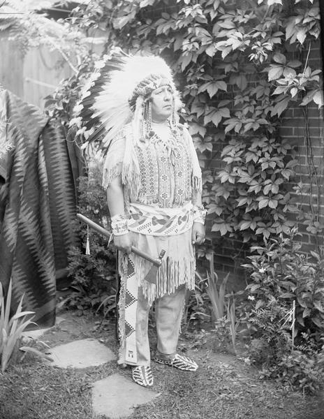 Full-length outdoor portrait of Ho-Chunk Chief Silver Tongue standing holding an axe. Brick wall, foliage, and blankets in the background.