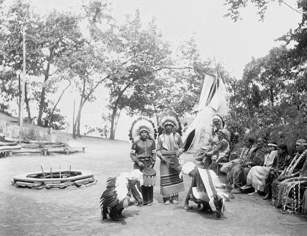 Ho-Chunk Eagle Dance. Two dancers crouch in front of two men with drums. Ho-Chunk men and women sit on a bench at right. Tepee in back.