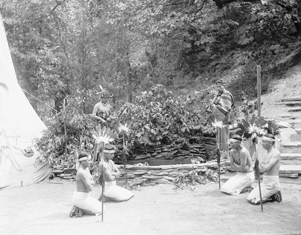 Ho-Chunk flute ceremony or prayer for rain. Four men kneel in a square next to poles bearing feather headdresses. A flute player and a man with a rattle stand behind them. Tepee at left.