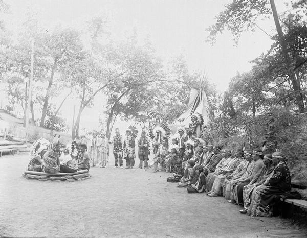 Group of Ho-Chunk at Stand Rock Amphitheater. Several men sit in a ring as other Ho-Chunk look on. Tepee in the background. This is where the Stand Rock Indian Ceremonial is performed.