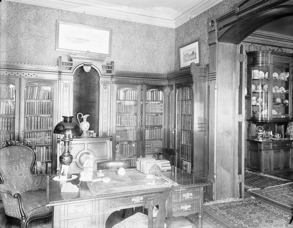 Interior view of Upham residence. There are bookshelves along the back wall. A large, arched doorway leads into another room on the right, with a glass-fronted cabinet displaying china.