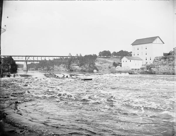 Several men on a raft going over the dam at Munger's Mill. There is a railroad bridge in background.