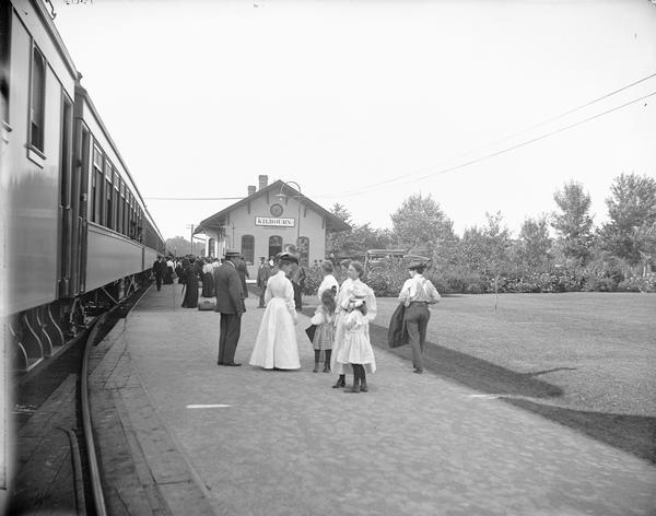 Kilbourn railroad station looking north. A crowd of people are standing on the platform. Carriages are parked behind the station building.