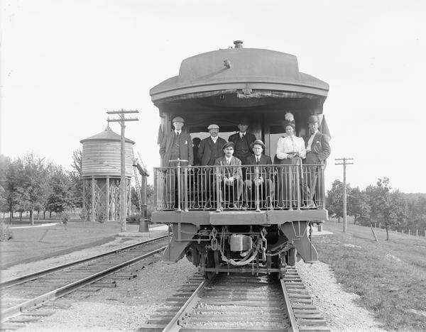 Group of people posing on the observation platform of a train at the Kilbourn station. There is a water tower on the left.