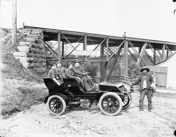 A.C. Bennett posing in a car, which is parked in front of Kilbourn Bridge, with three other men. A man is standing at the front of the car and another is sitting on the bridge.