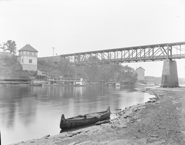 View from shoreline, with a canoe in the foreground. Across the river on the opposite shoreline is the "Apollo No. 1" steamboat at Dells Landing, at Kilbourn. There is another steamboat on the right under the bridge. Munger's Mill is beyond the bridge.