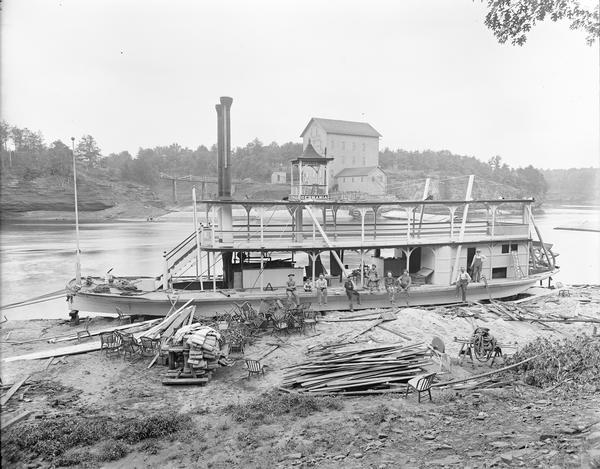View down hill of the "Germania" steamboat along the shoreline in the foreground, across the river from bridge and Munger's Mill, which are in the background. Several men and a boy are sitting on the edge of the "Germania." Chairs, lumber and miscellaneous items are piled on the shore in front of the "Germania."