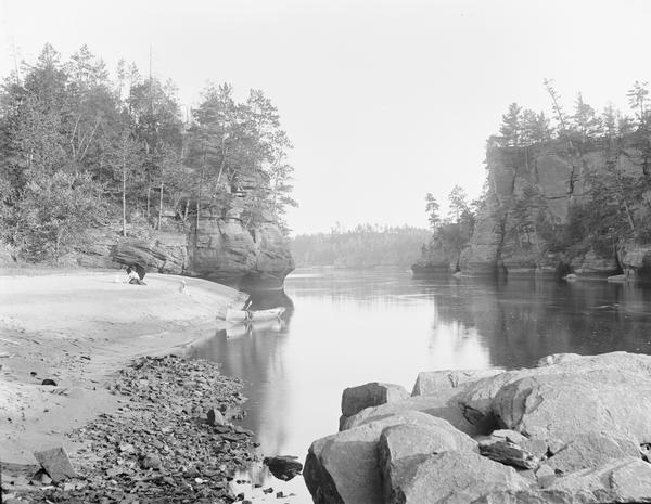 View along shoreline towards a woman and two children sitting on the sandy shore. There is a canoe at the edge of river.