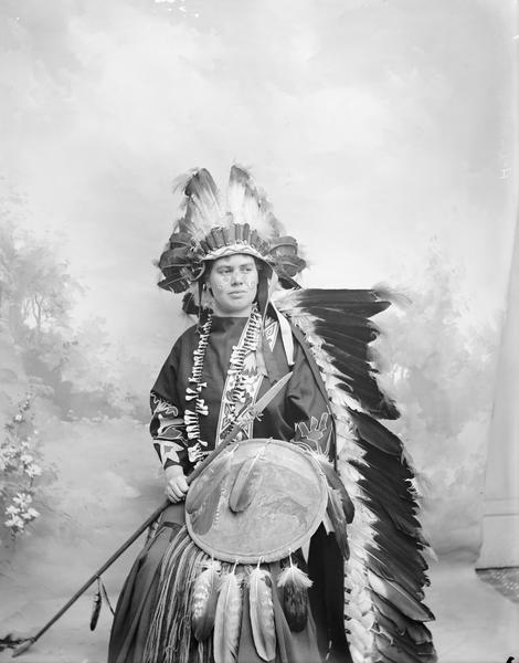 Studio portrait in front of a painted backdrop of an unidentified person wearing Native American costume. Subject is sitting and is holding a spear and a shield.