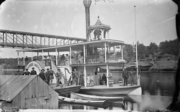 View from shoreline of passengers posed on steamboat <i>Alexander Mitchell</i> with a bridge and opposite shoreline in the background. One man leans on a harp. Rowboats are in front of the steamboat near what appears to be a boathouse.