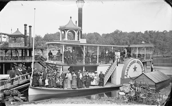 Steamboat <i>Alexander Mitchell</i> with passengers posed standing. Chairs are piled on shore at right. Edge of steamboat <i>Dell Queen</i> at left. Bridge in background.