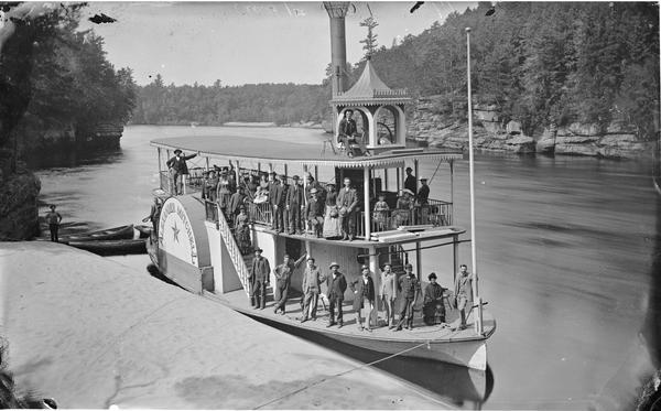 View from shoreline of the steamboat <i>Alexander Mitchell</i> docked at edge of river with passengers posed on board.