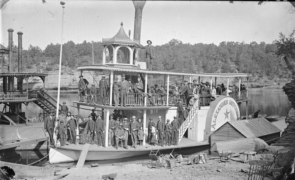 Steamboat <i>Alexander Mitchell</i> with passengers posing on board. Man with harp on bow. Edge of "Dell Queen" on left. Chairs piled on shore.