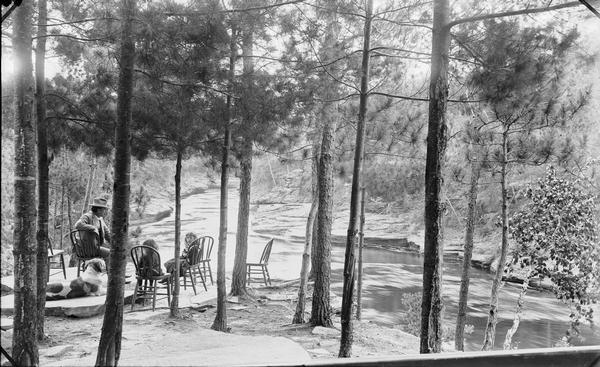 Larks Hotel, downstream. A man (George Crandall) and two children (his daughters) are sitting in chairs under trees overlooking the Wisconsin River, with the rapids in the background. There is a dog sitting on the ground.