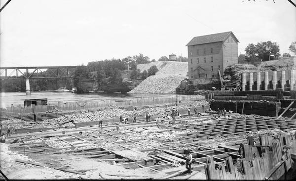 Elevated view of Kilbourn Dam, building apron. Men are working on the dam, and a bridge is in the background.