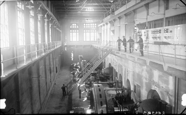 Interior view from balcony of Kilbourn Dam Powerhouse. Four men stand at the balcony railing near stairs that lead to the ground floor. Another man stands at the bottom of the stairs near the generators.