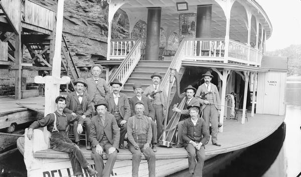 H.H. Bennett and men posing on bow of "Dell Queen" steamboat. There are two men with violins, one with a harp.