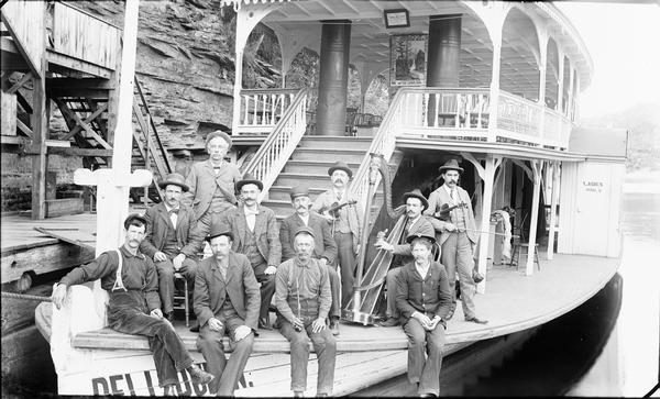 H.H. Bennett and group of men posing on bow of "Dell Queen" steamboat. There are two men with violins, one with a harp.
