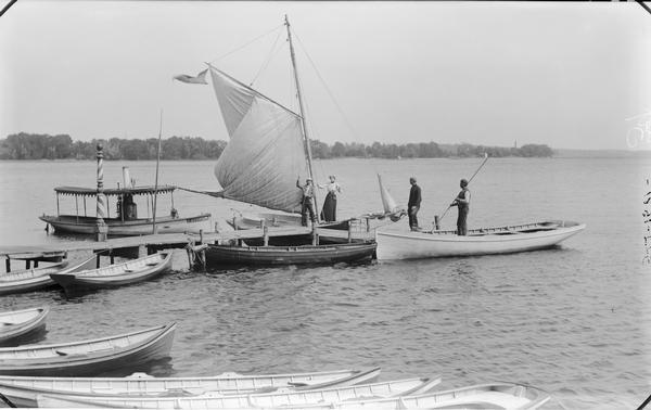 View from shoreline towards boats at a dock in the Fox Lake Area. A man and woman are standing in a sailboat, another man is standing on the dock, and a another man is standing in a boat. A row of boats are lined up along the shoreline in the foreground.