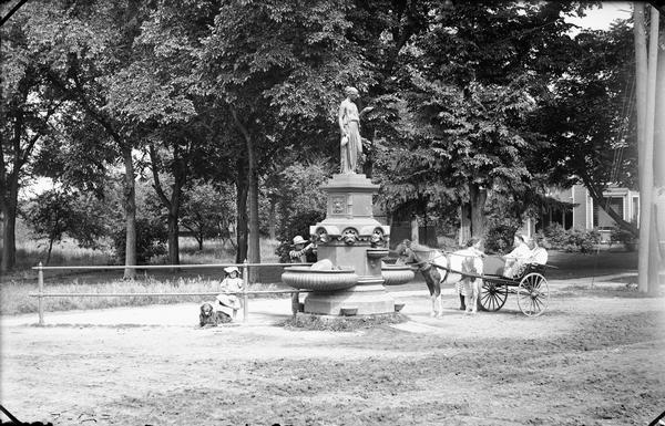 Children at fountain on Broadway and Superior. Two girls are sitting in a pony-cart, a girl is looking through a railing with a dog, while a boy is filling a vessel from the fountain.