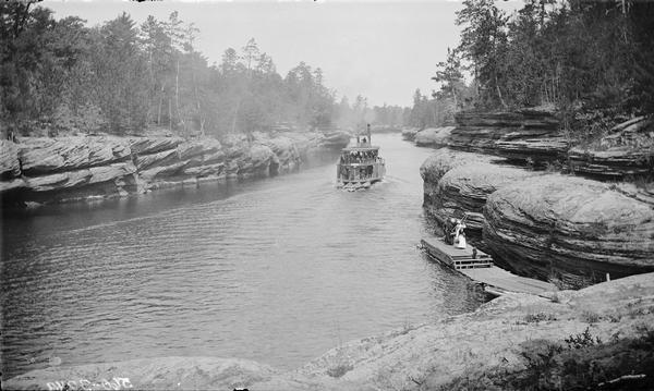 Elevated view from rock of Eaton Grotto. On the river is a steamboat with passengers. People are standing on a dock at river's edge.