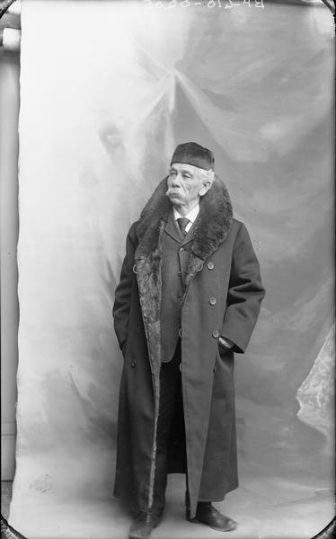Full-length studio portrait of H.H. Bennett, who is standing and wearing a long coat with a fur collar.