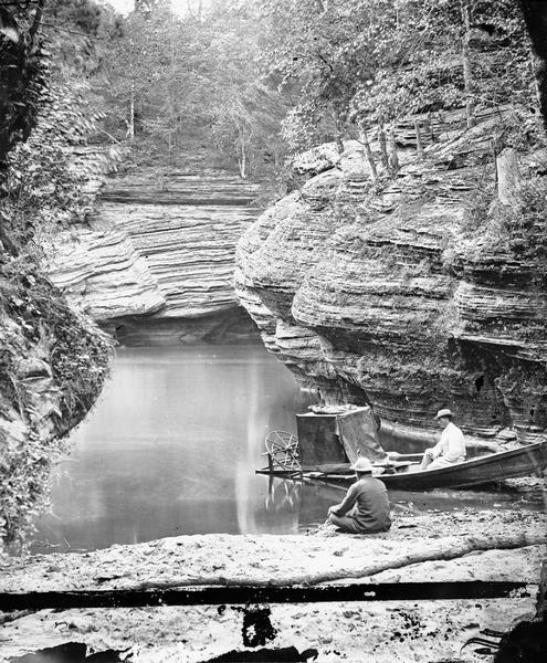 Boat and dark tent at Gates Ravine. One man is sitting in a boat on the water. Another man is in the foreground sitting on the rock face near the shoreline.