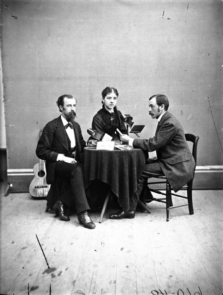 Studio portrait of Chet and Ella Weber, and Holly Weber sitting around a table and looking at stereograph cards. There is a guitar leaning against the wall in the background.
