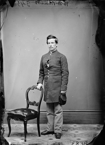 Full-length studio portrait of H.H. Bennett standing. He is wearing a uniform, holding a hat in his left hand, and resting his right hand on the back of a chair.